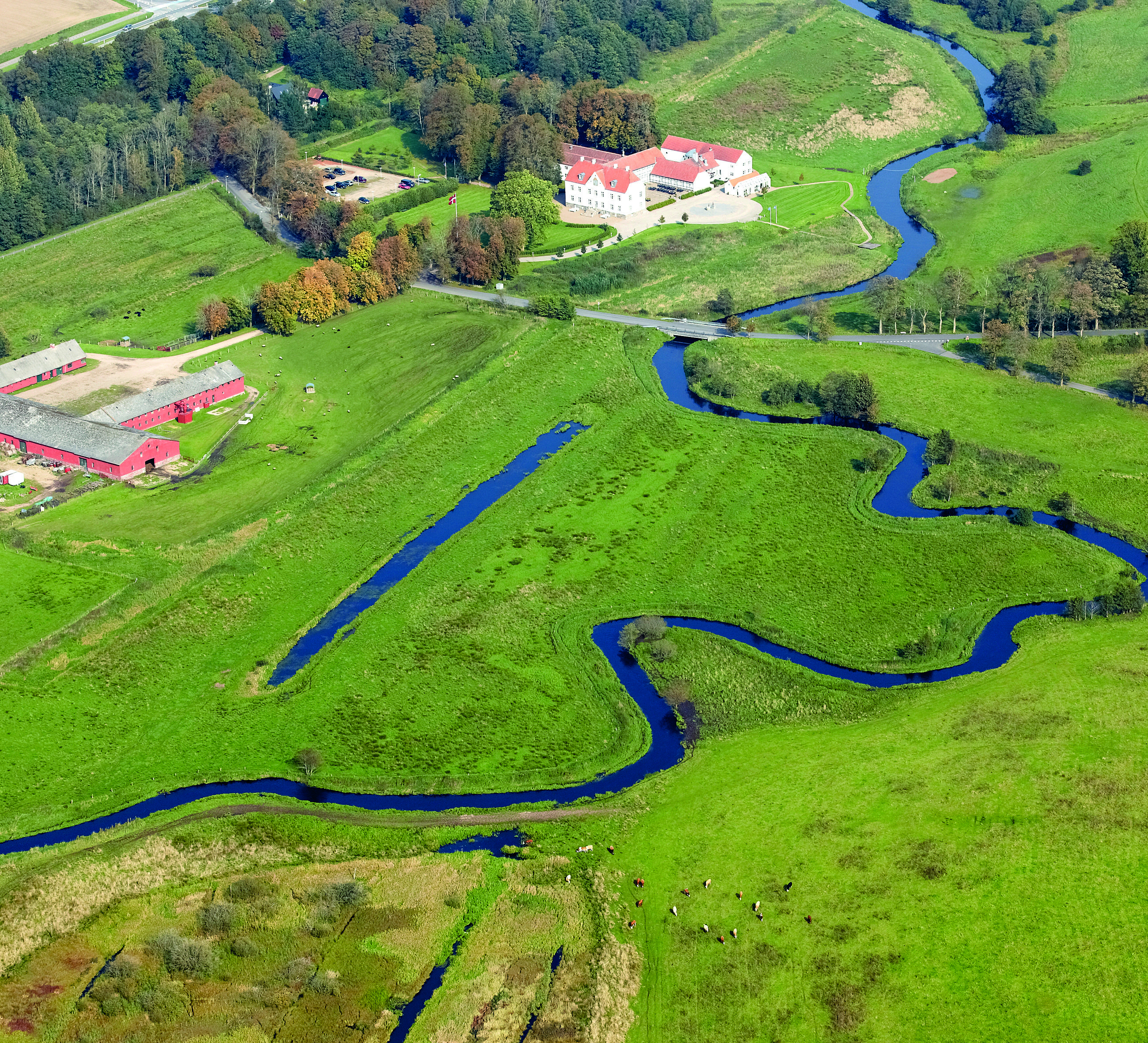 Luftfoto over vådområde ved Vejle Ådal med Vejle å, der snor sig igennem. På billedet ses en gård og Hotel Haraldskær.